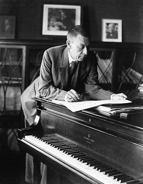 Sergei Rachmaninoff leaning over a piano reading a newspaper